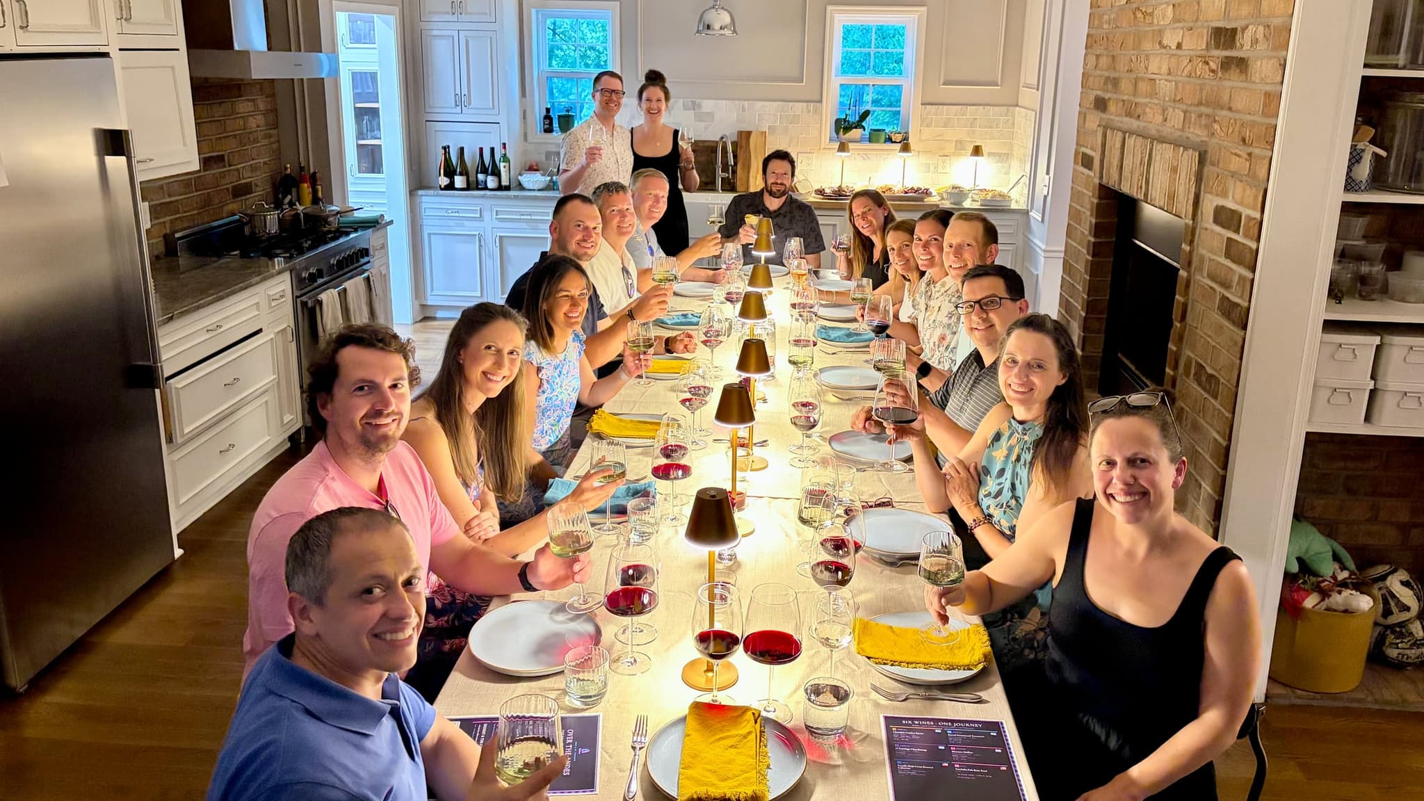 Fifteen guests raise their glasses down one long table, tasting placemats and flights of wine at every seat, the two hosts standing at the back of the room