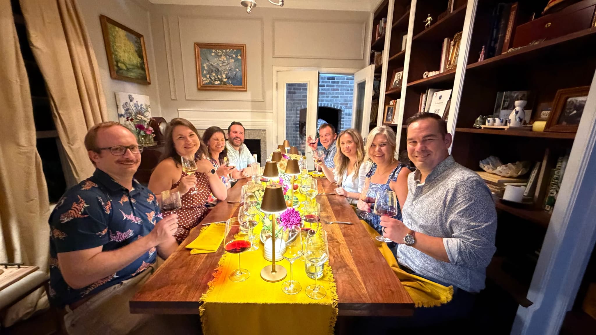 Eight people seated down a long wooden table with a mustard-yellow runner raise their wine glasses, small brass table lamps and magenta flowers down the middle, bookshelves along one wall and a fireplace at the far end